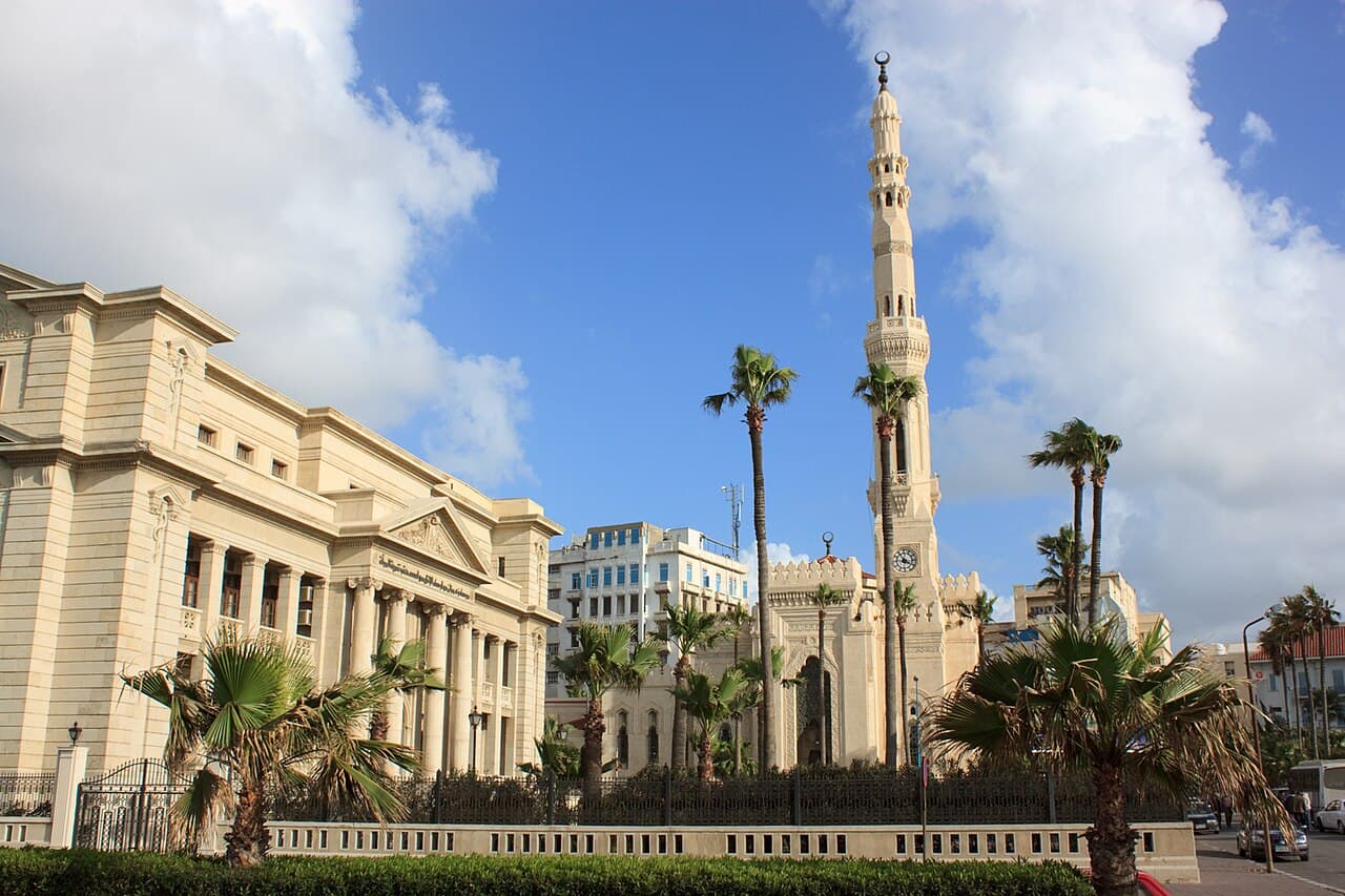 مئذنة مسجد القائد إبراهيم ترتفع فوق النخيل، وإلى جوارها مبنى كلاسيكي بأعمدته