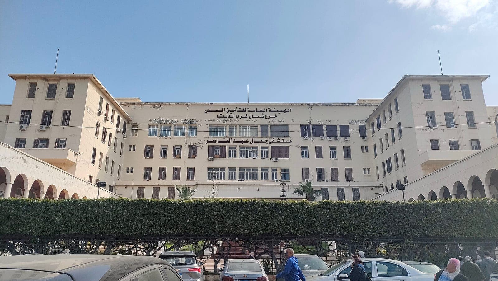 المبنى الرئيسي لمستشفى جمال عبد الناصر بالإسكندرية خلف صف من الأشجار المشذبة وساحة انتظار سيارات.